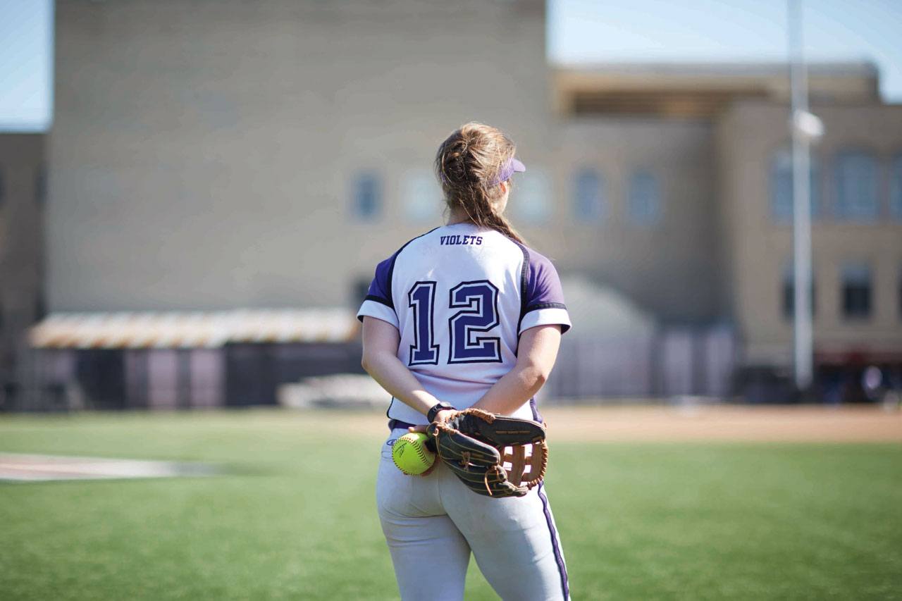 A member of the NYU Softball team stands on the field with her back to the camera.
