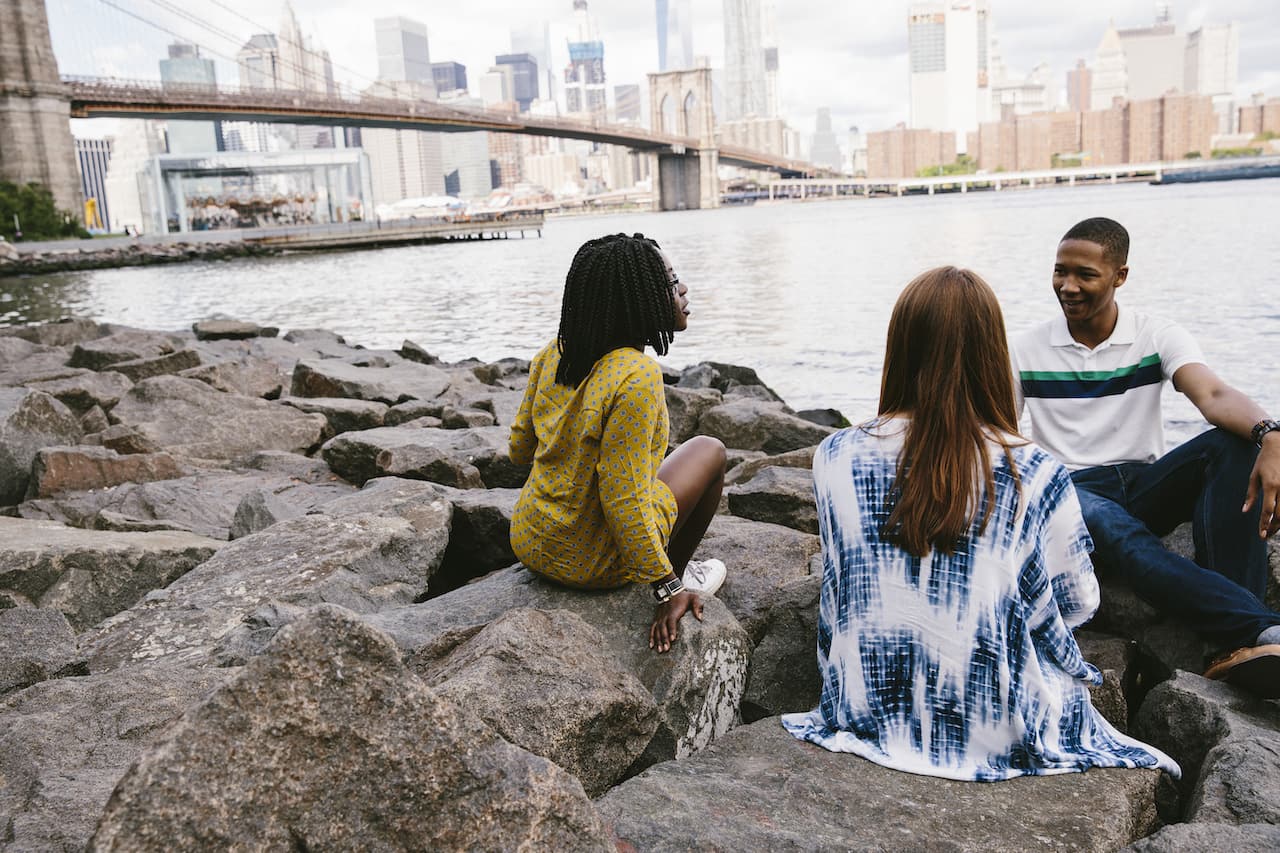 [Three students sit and talk along the river whith a view of the Brooklyn Bridge and downtown NYC behind them.]