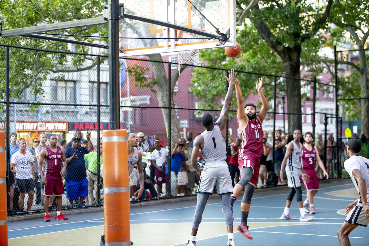 [A basketball player goes up for a jump shot while being guarded by the other team. The teams play on an outdoor court with lots of people watching from behind a fence.]]