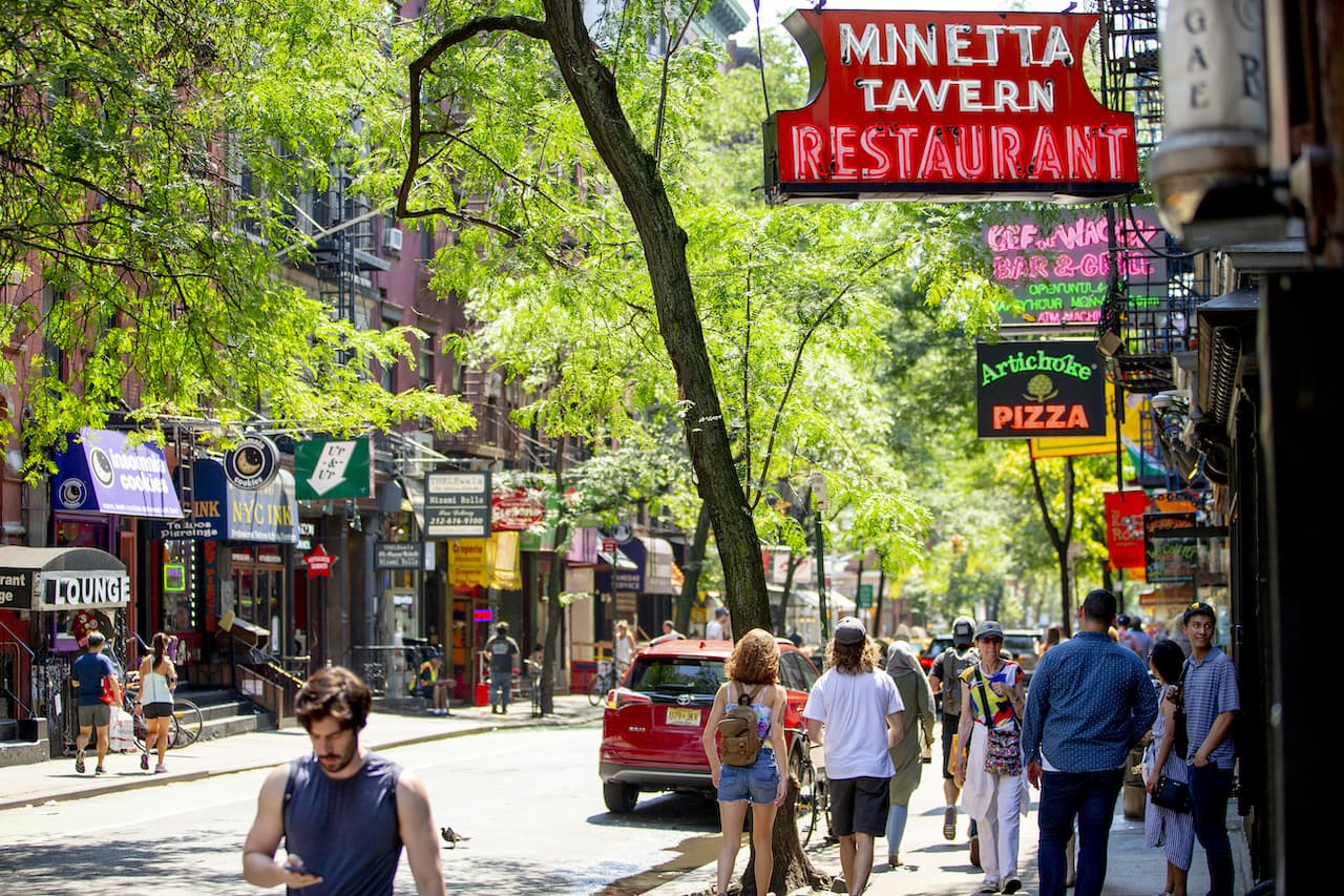 [People walk down a busy street in Greenwich Village, passing by lit up signs or restaurants, bars, and stores.]