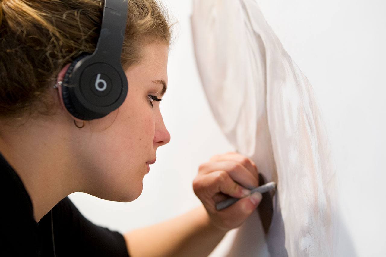 A student wearing headphones leans in close to touch up her art with an oil stick