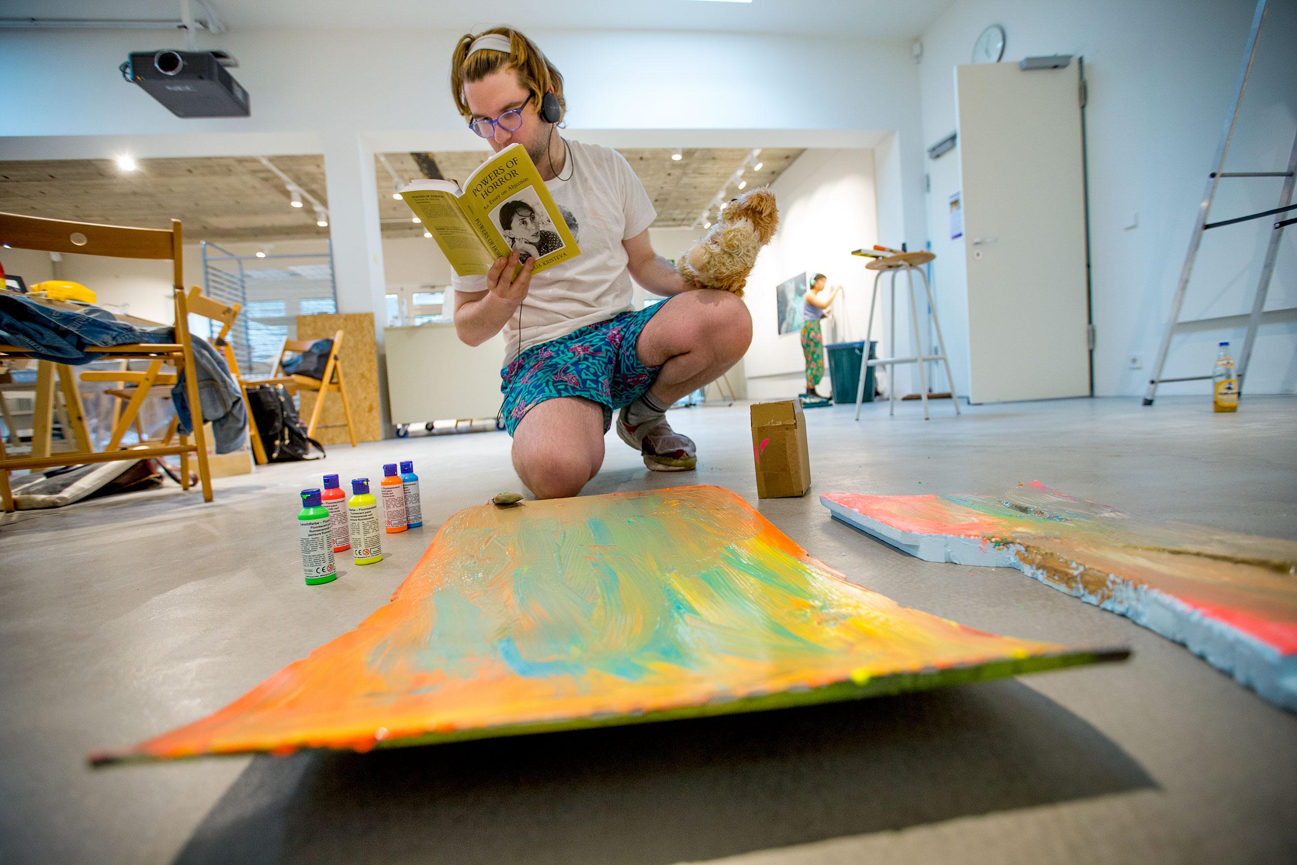 A student studying art abroad reads from a book while crouched over his painting on the floor