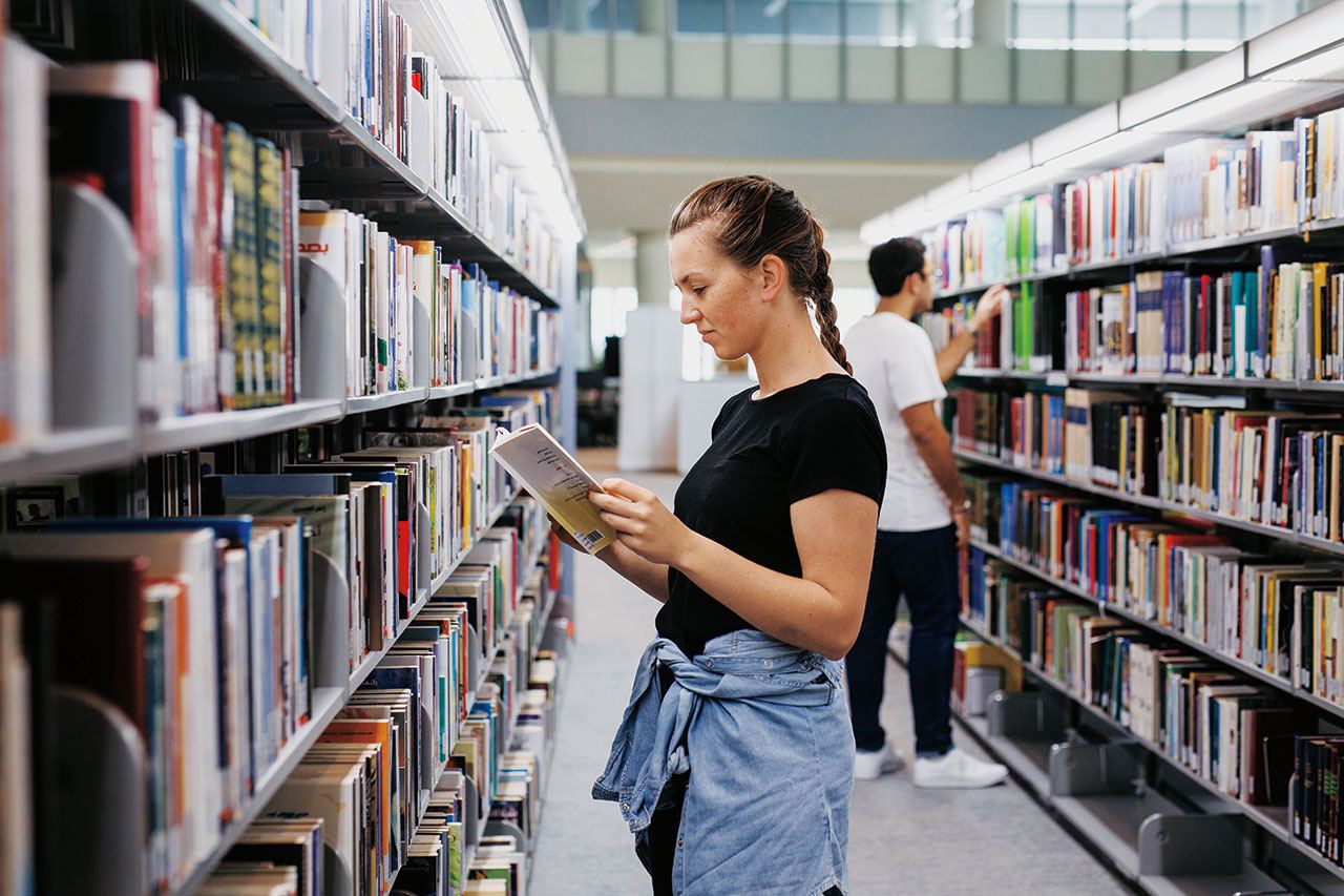 A female student reading a book in the library stacks.