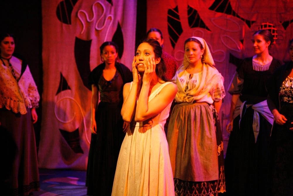 Students on a stage performing in costume. The woman in the forefront is wearing a dress and has her hands pressed to her cheeks.
