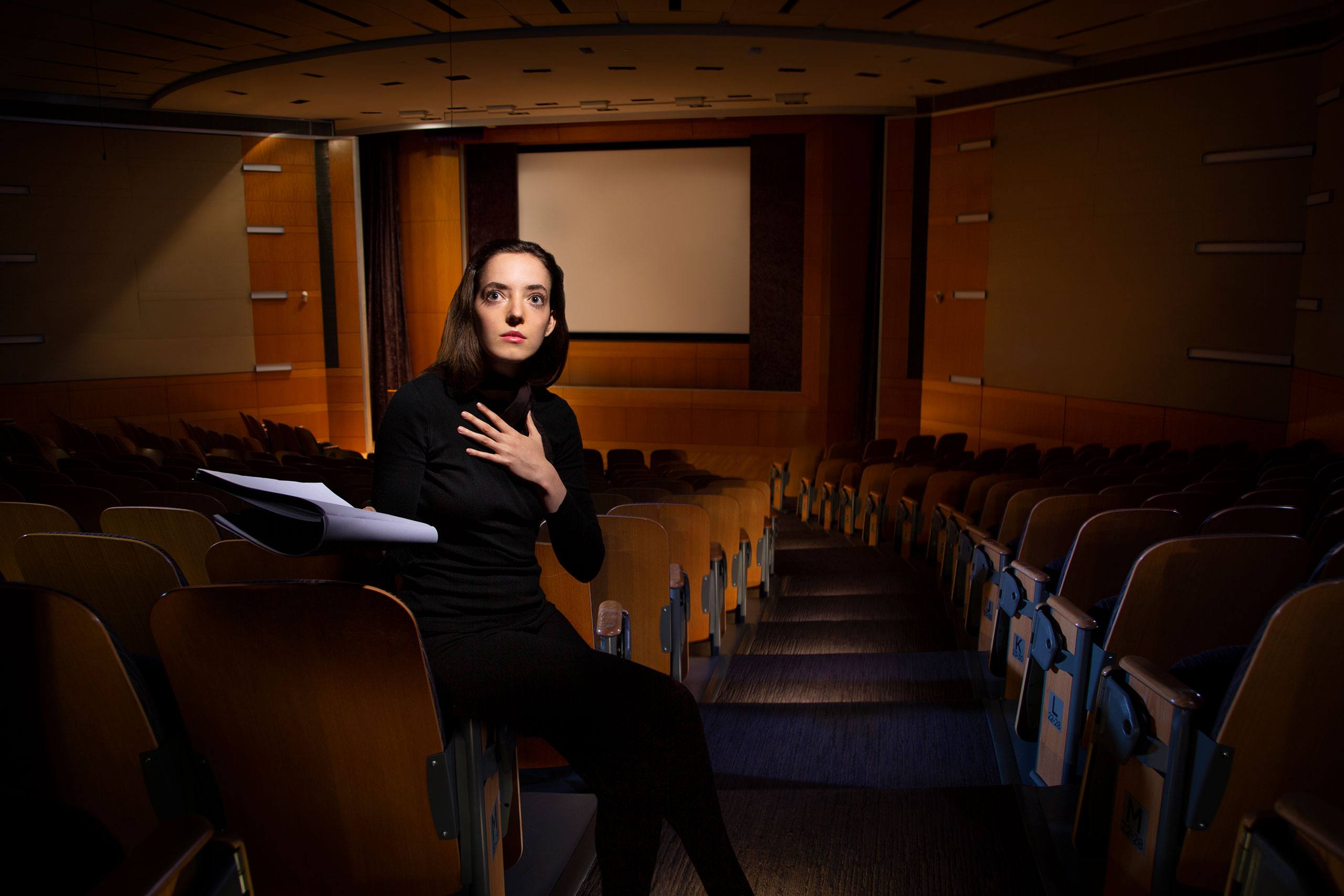 Student Adriana Virgili Alovisetti sits in an empty theatre looking upward and gesturing forward with a script in her hand.