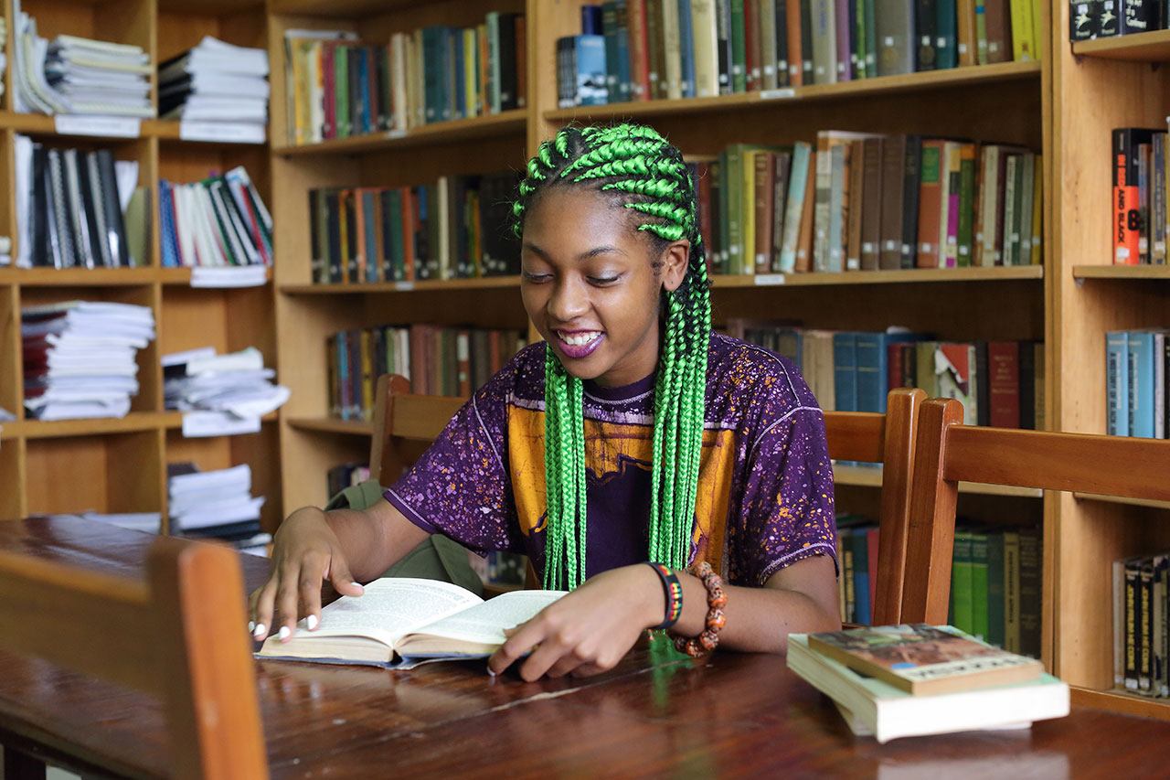 A Black student sitting in a library, reading a book, and smiling.