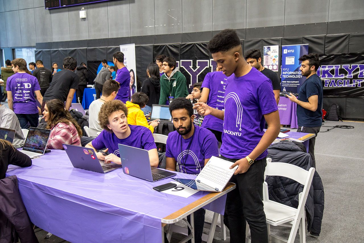 Three students, who are wearing purple-branded NYU shirts in support of the event, at HackNYU. Two students sit with computers in front of them and the third student stands and holds his laptop. Around them are other students who are also competing.