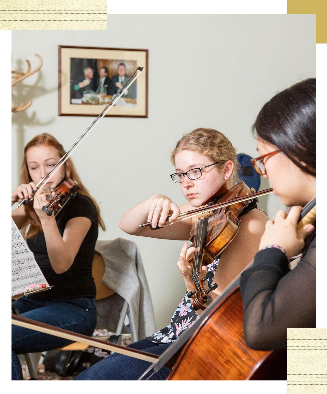 Three students playing instruments. Two playing the violin, while the third plays the cello.