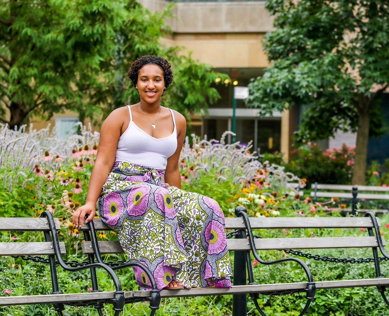 Student Mickayla Coutinho Brandao sitting on the top of a park bench, surrounded by an array of flora.