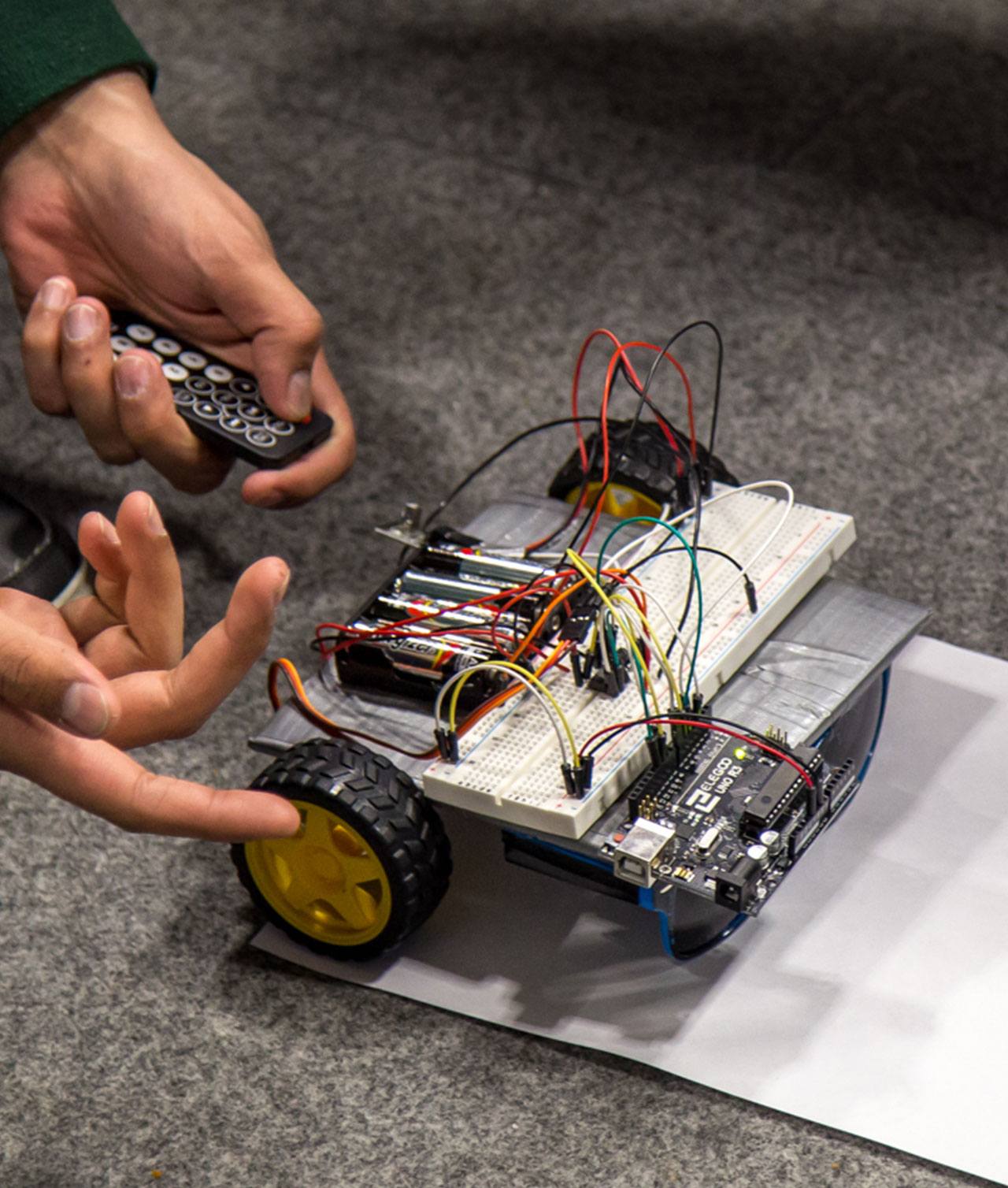 A close-up of a robotic creation and two hands. One hand holds a controller and the other gestures to the robot.