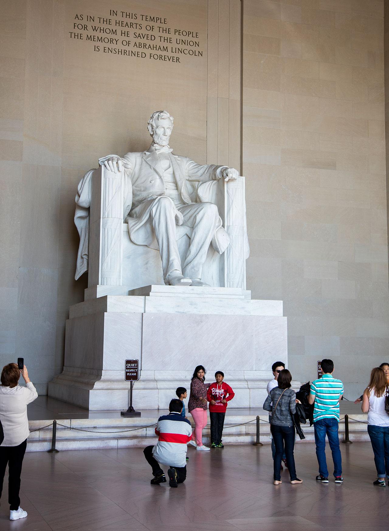 A statue of Abraham Lincoln at the Lincoln Memorial. People walk and take pictures around it.
