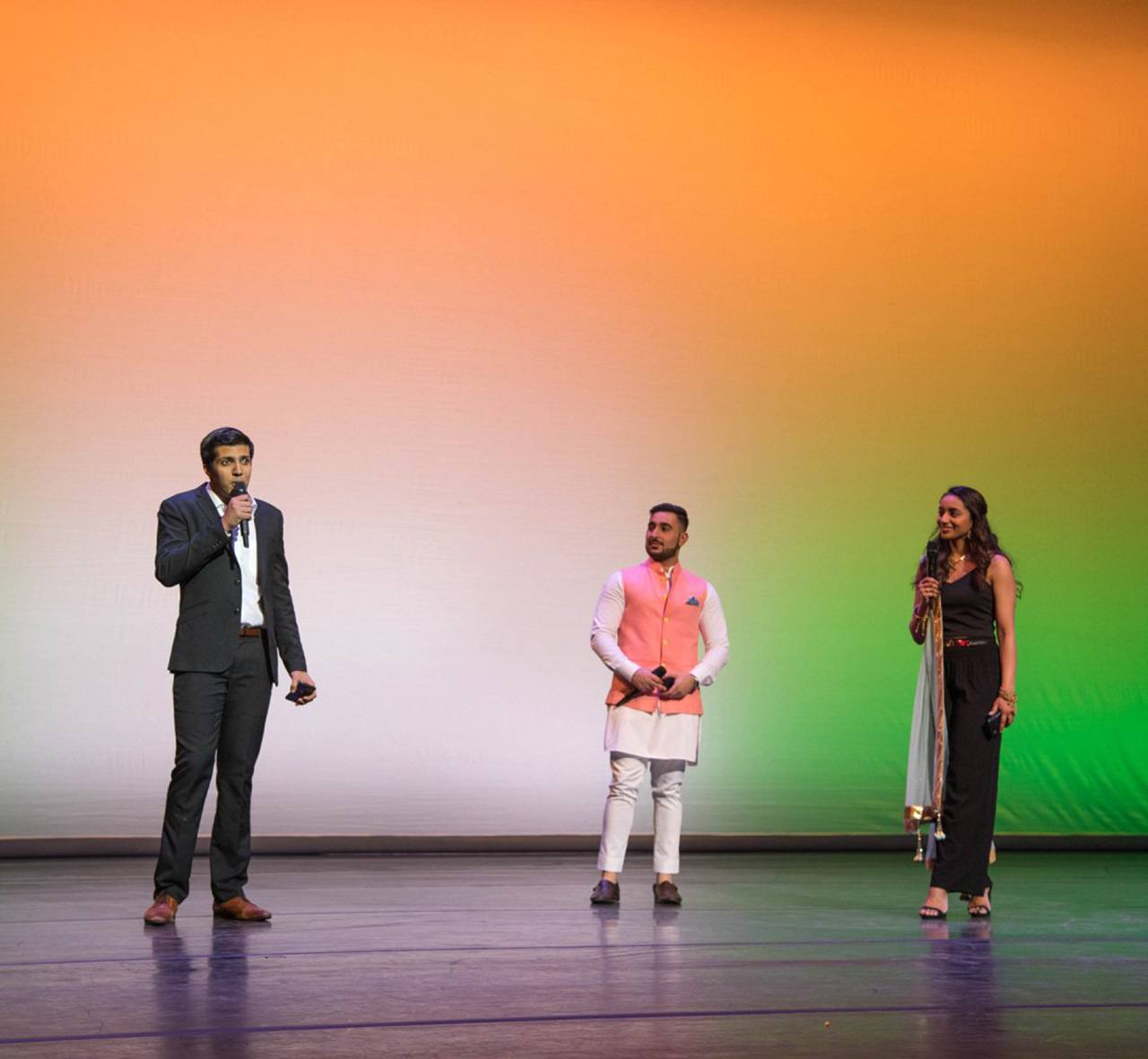Three students on stage holding microphones and standing in front of a decorative gradient background.