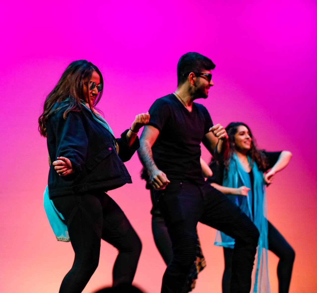 Students dancing on stage in Monsoon event. They are wearing dark clothing, sunglasses, and bright blue scarves.