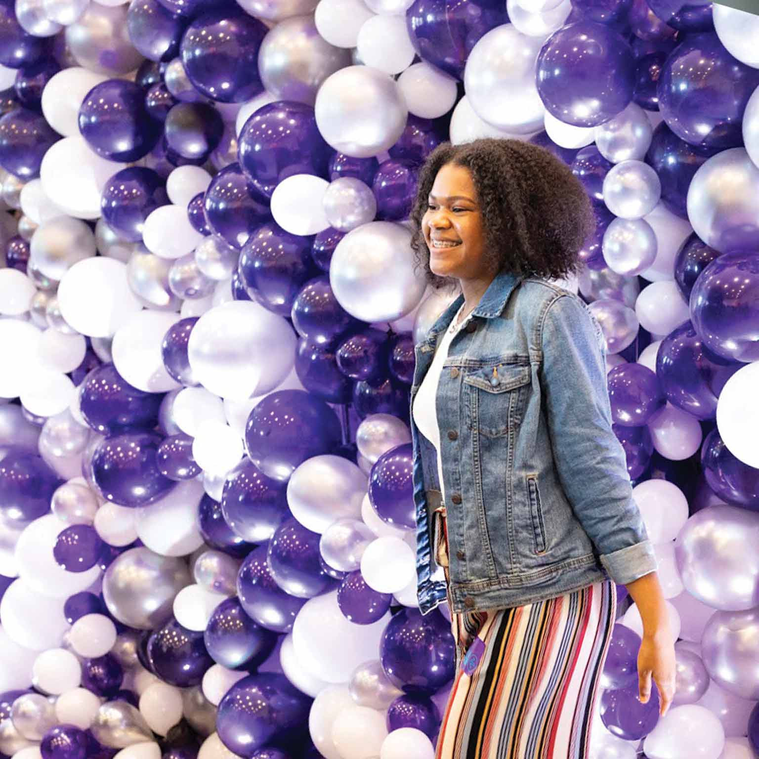 An NYU admitted student standing by a violet and white balloon wall.