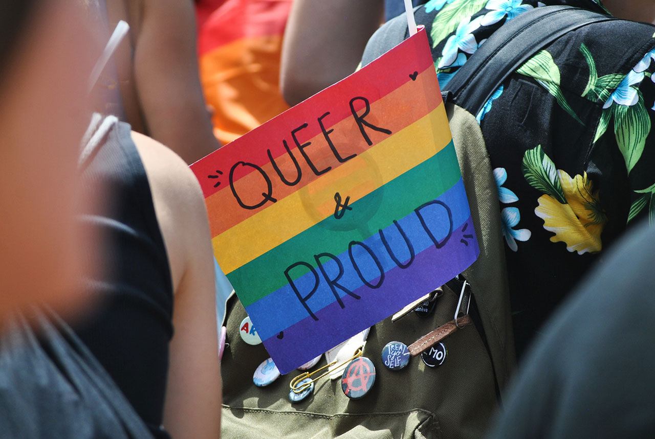 A flag that reads “Queer & Proud” jutting out of a backpack.