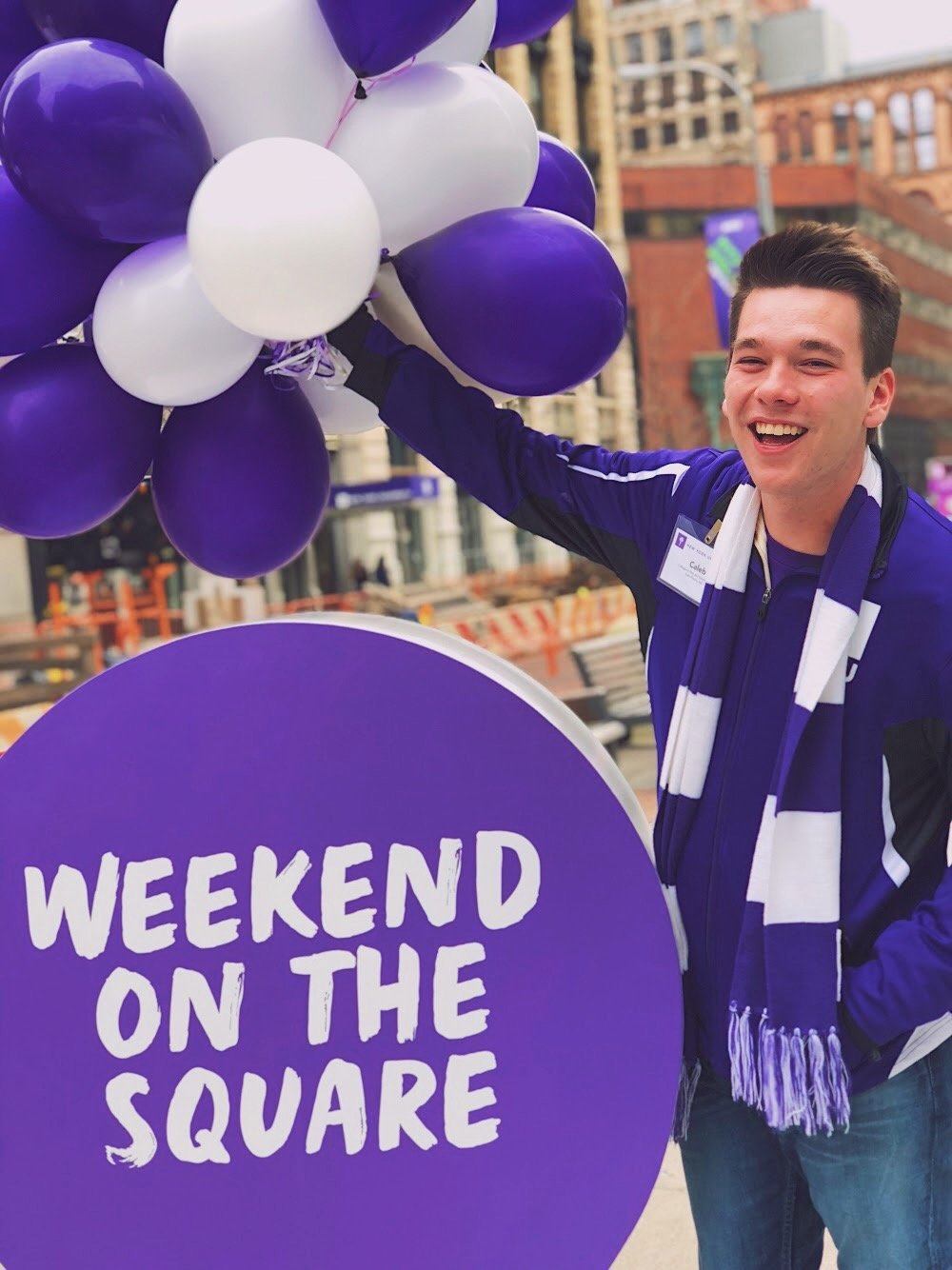 NYU student Caleb Martin helps students choosing NYU while wearing NYU attire and holding purple and white balloons.