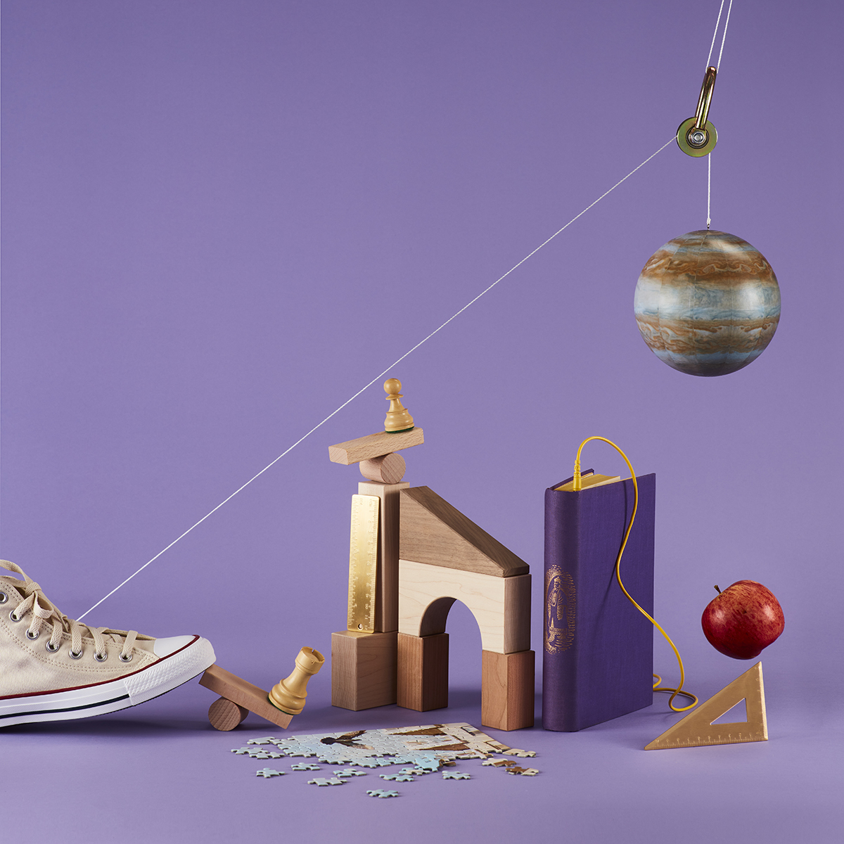 Still life of a white Converse shoe kicking off a Rube Goldberg machine involving chess pieces, wooden bloods, a book, an unmade puzzle, a planet on a pulley, and an apple rolling down a triangle.