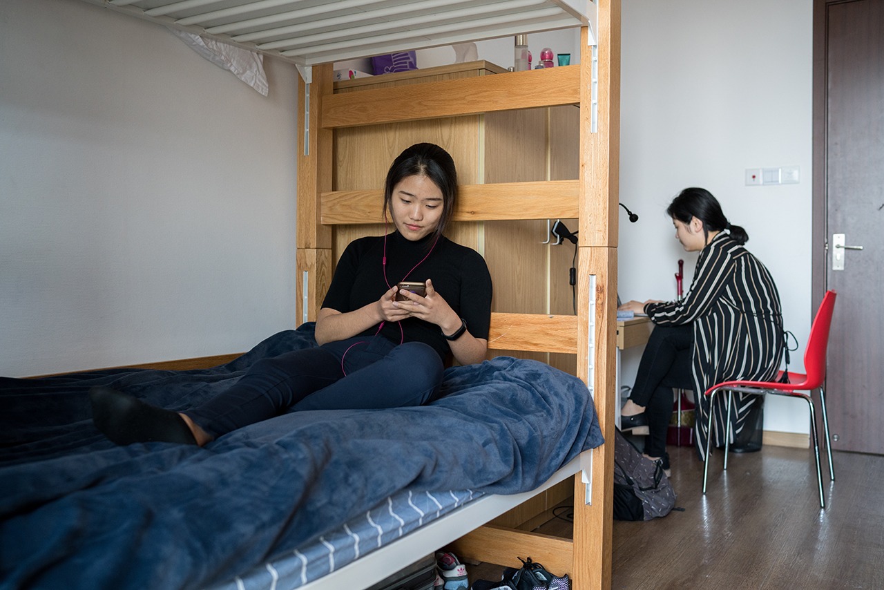 A student sitting in a dorm room bed, her roommate is sitting in a desk behind her.