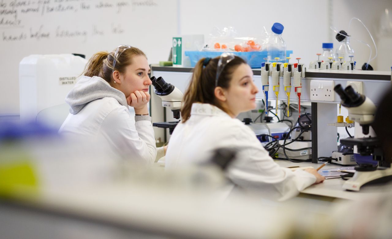 Two students sitting in a science lab wearing lab coats.