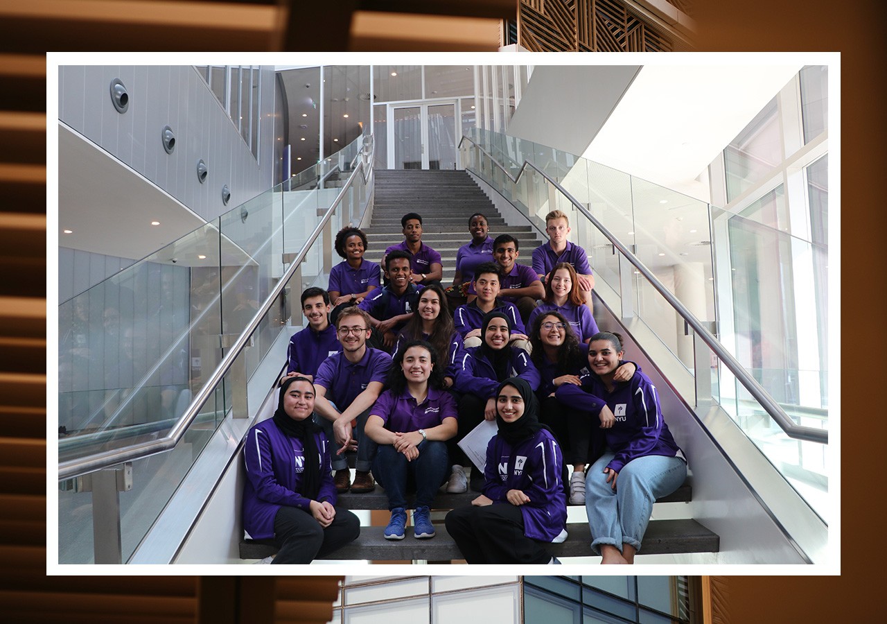 NYU students posing for a group photo on a set of stairs. The students are wearing NYU tee shirts and sweatshirts.