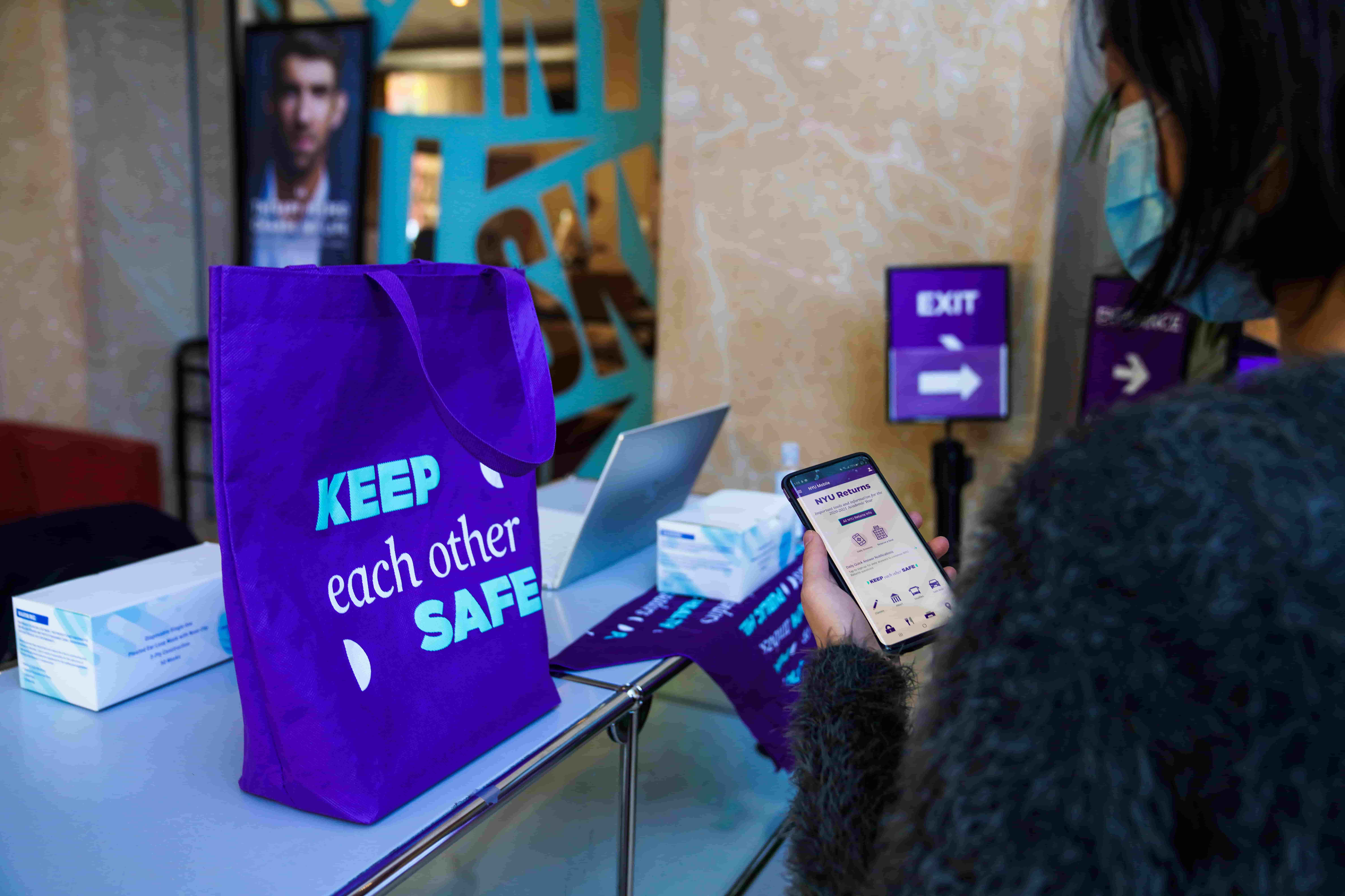 An NYU student using the daily screener and a tote bag that reads “keep each other safe.