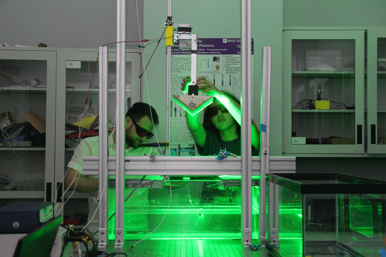 Two students working with lab equipment. The machine they are working with is emitting a green glow, and the students are wearing protecting googles
