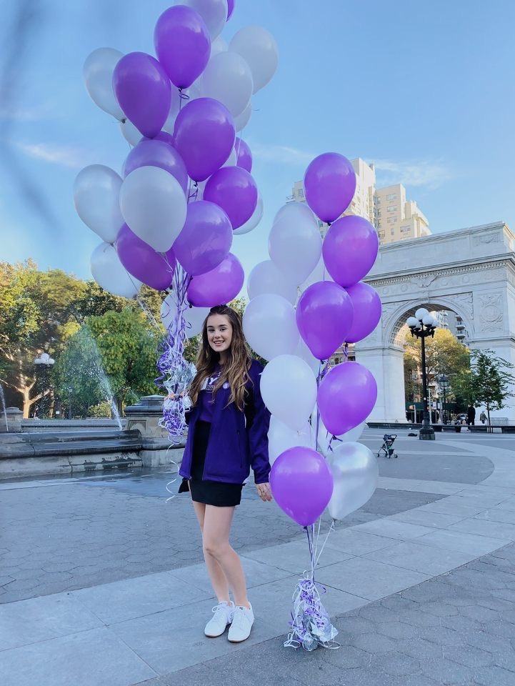An NYU Admissions Ambassador holding white and violet balloons in Washington Square Park.