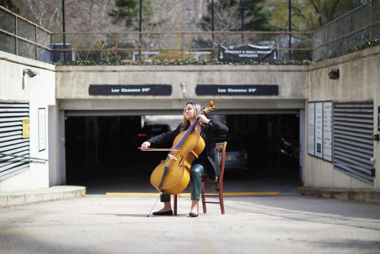 A girl playing a cello in front of a parking garage.