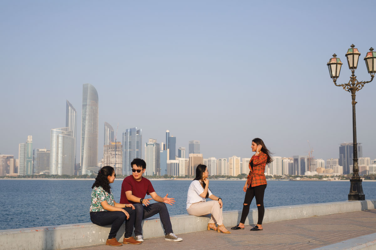 A group of students sitting near a large body of water with the Abu Dhabi skyline in the distance.