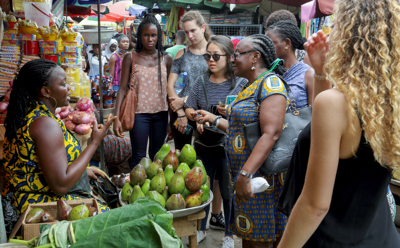 A large group of students and a guide interacting with a shop owner in Accra.