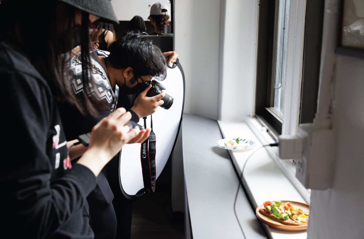 A student with a digital camera taking a photo of a food dish presented on a windowsill.