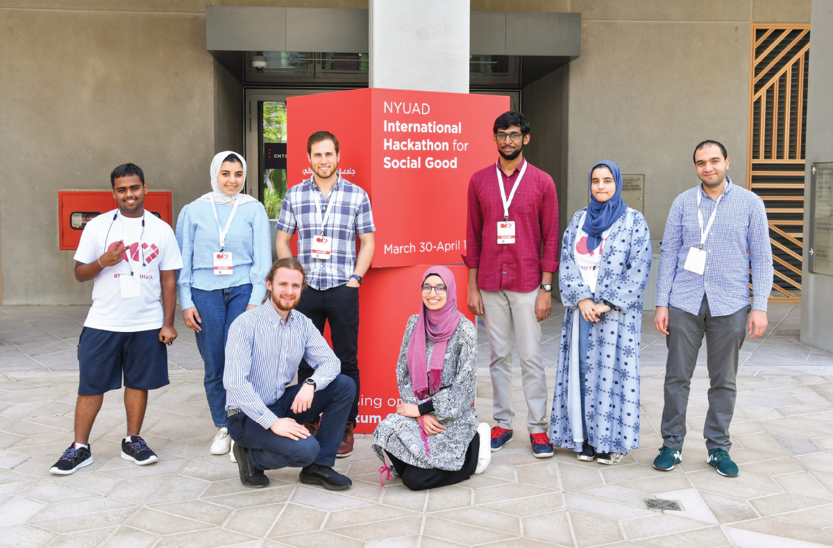 A group of students in front of a red sign that reads, “NYUAD International Hackathon for Social Good.”