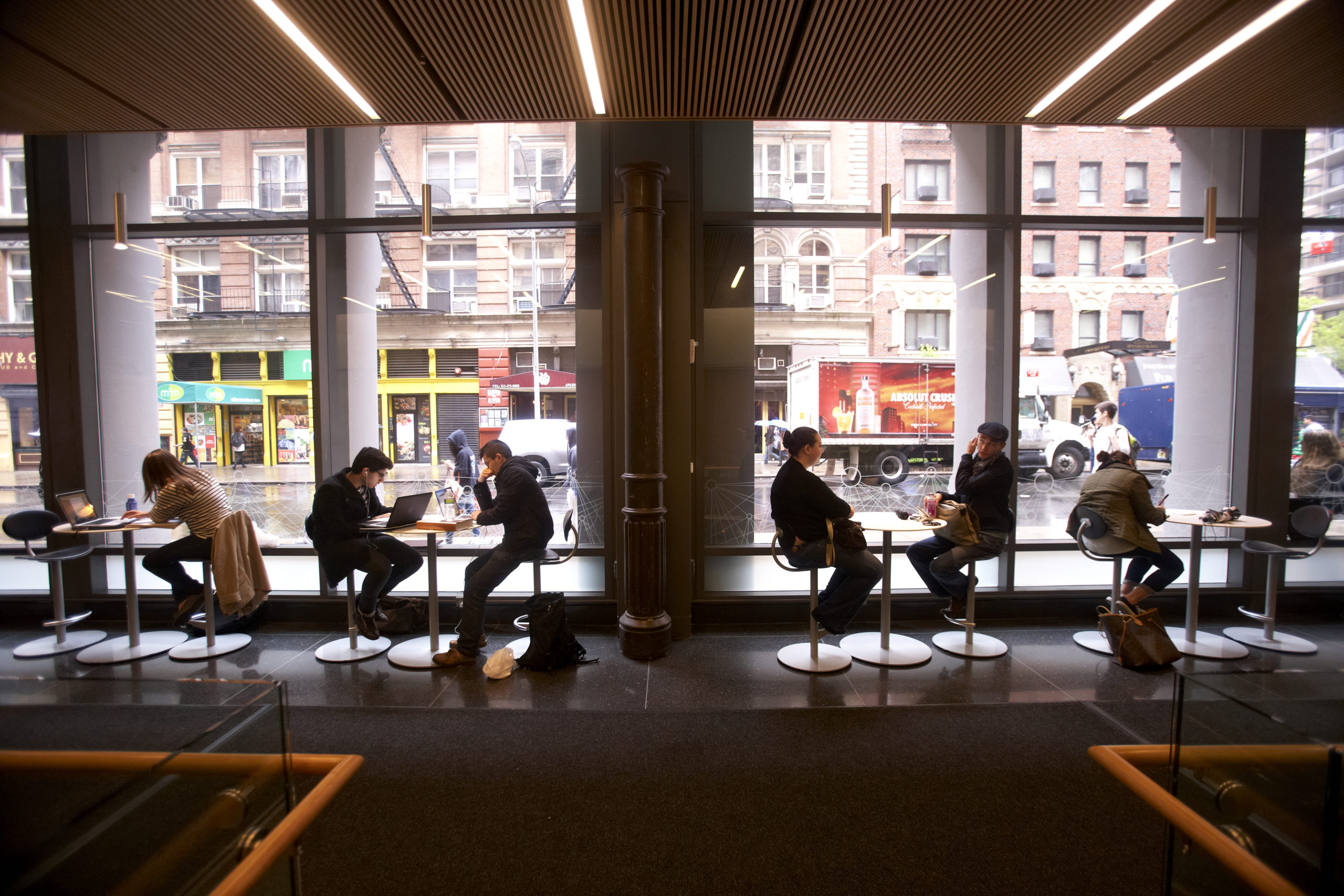 Students sitting at high tables in a lounge area on NYU’s campus. The room has high windows and have a view of the street.