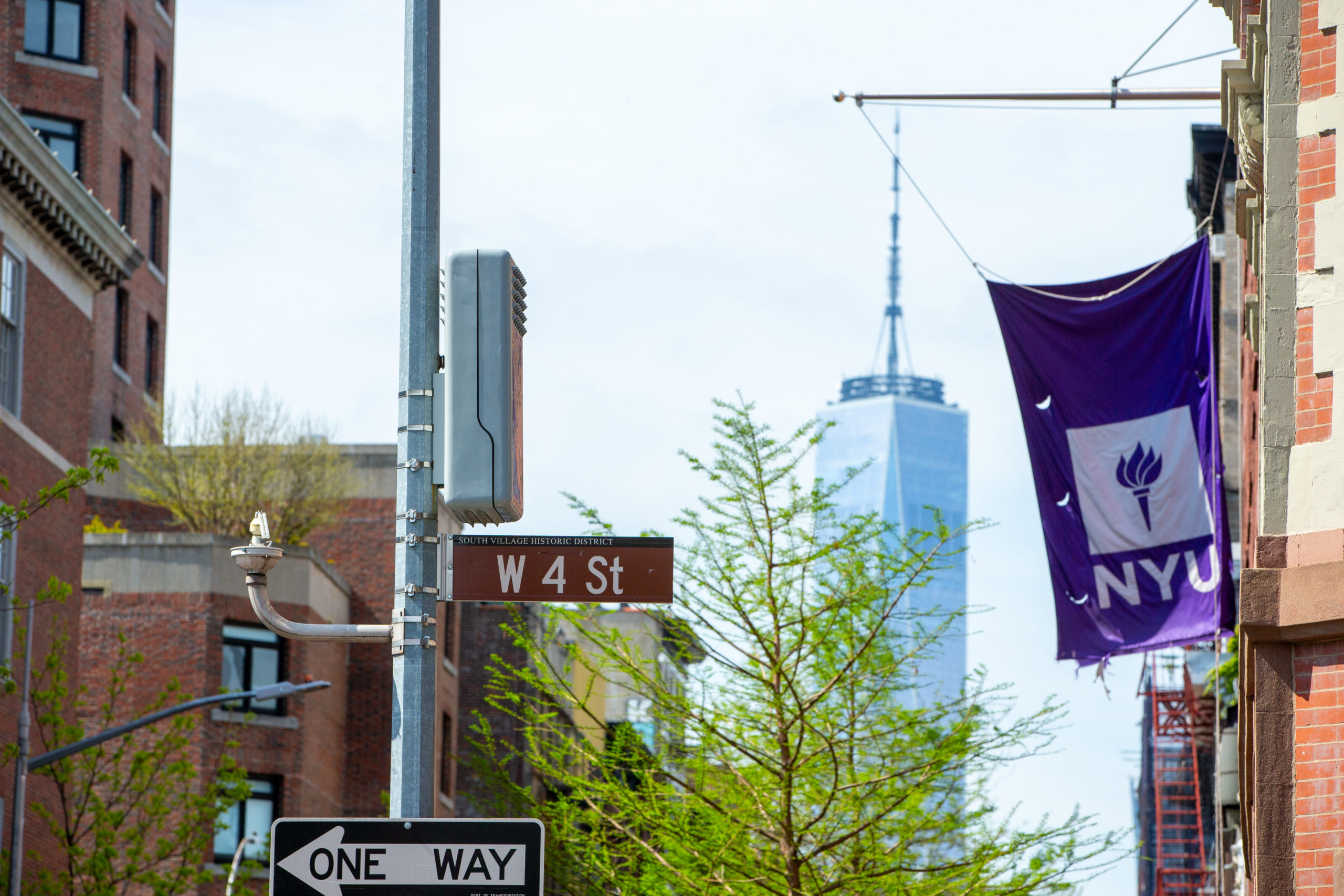 An NYU flag attached to a building. A sign for West Fourth Street is in front of it, and the New York City skyline with the World Trade Center is behind it.