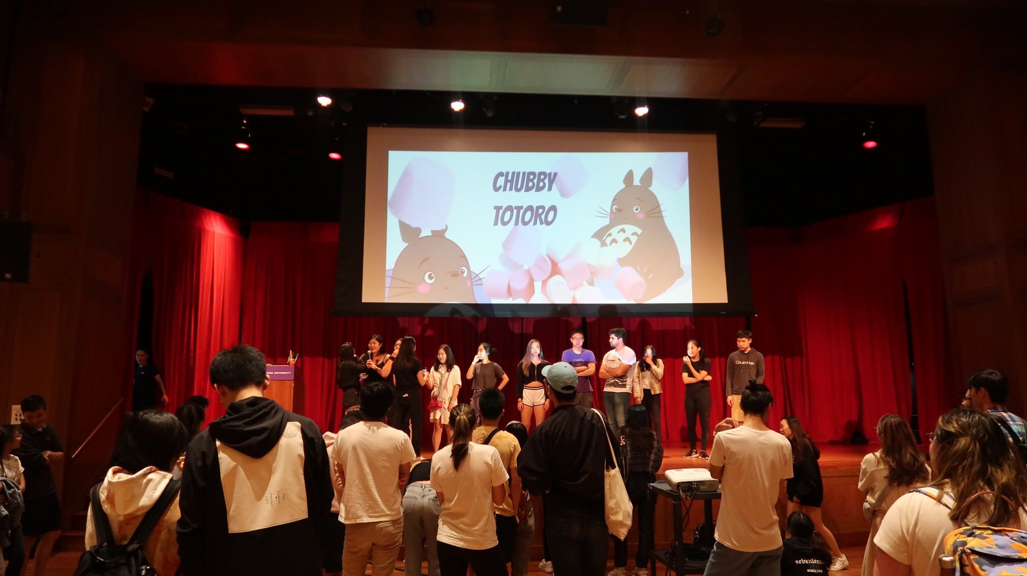 An image titled Chubby Totoro is projected behind students standing onstage during a game.
