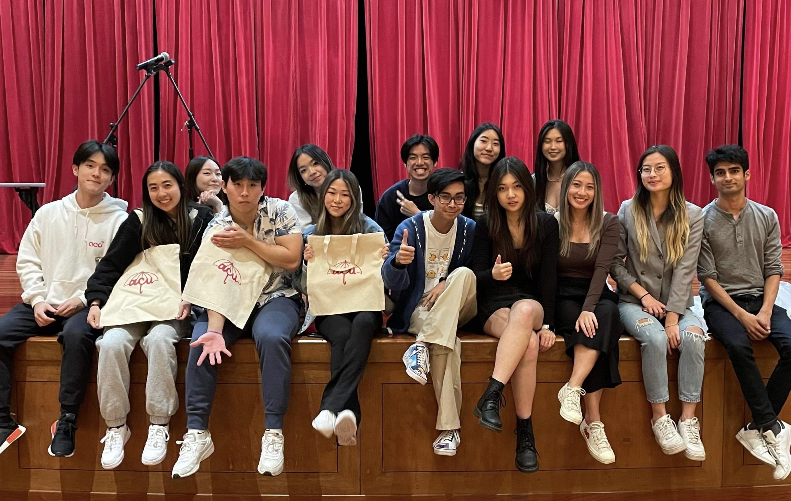 Students sitting on a stage and smiling with ACU tote bags.