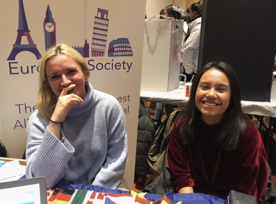 Two European Society students sitting and smiling at a table covered in European Flags. A “European Society” sign is behind them.