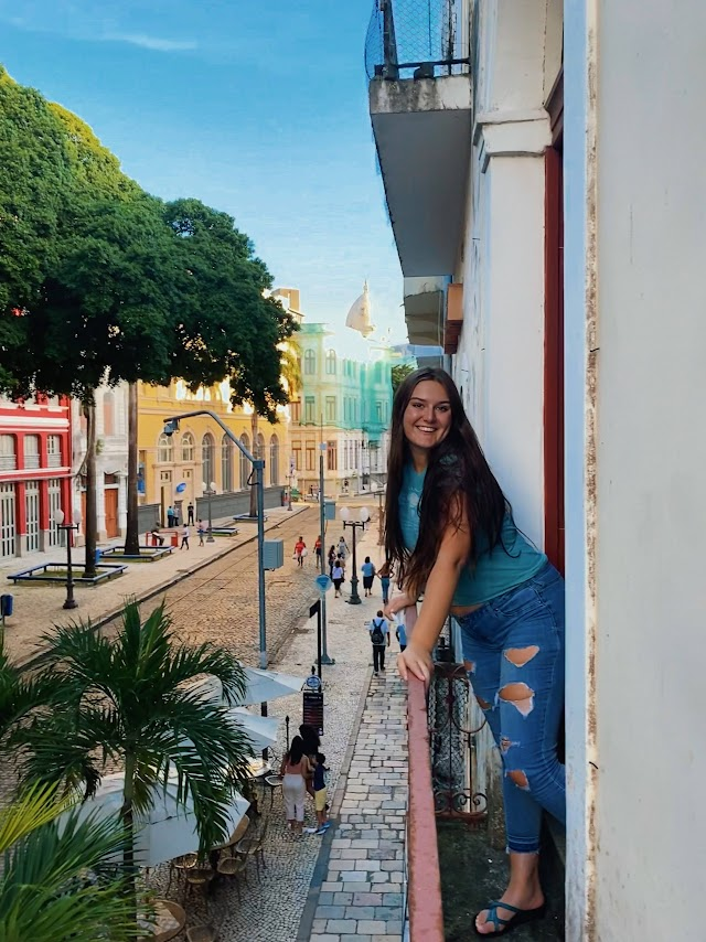 Sophia standing on a balcony with colorful buildings and trees in the background.
