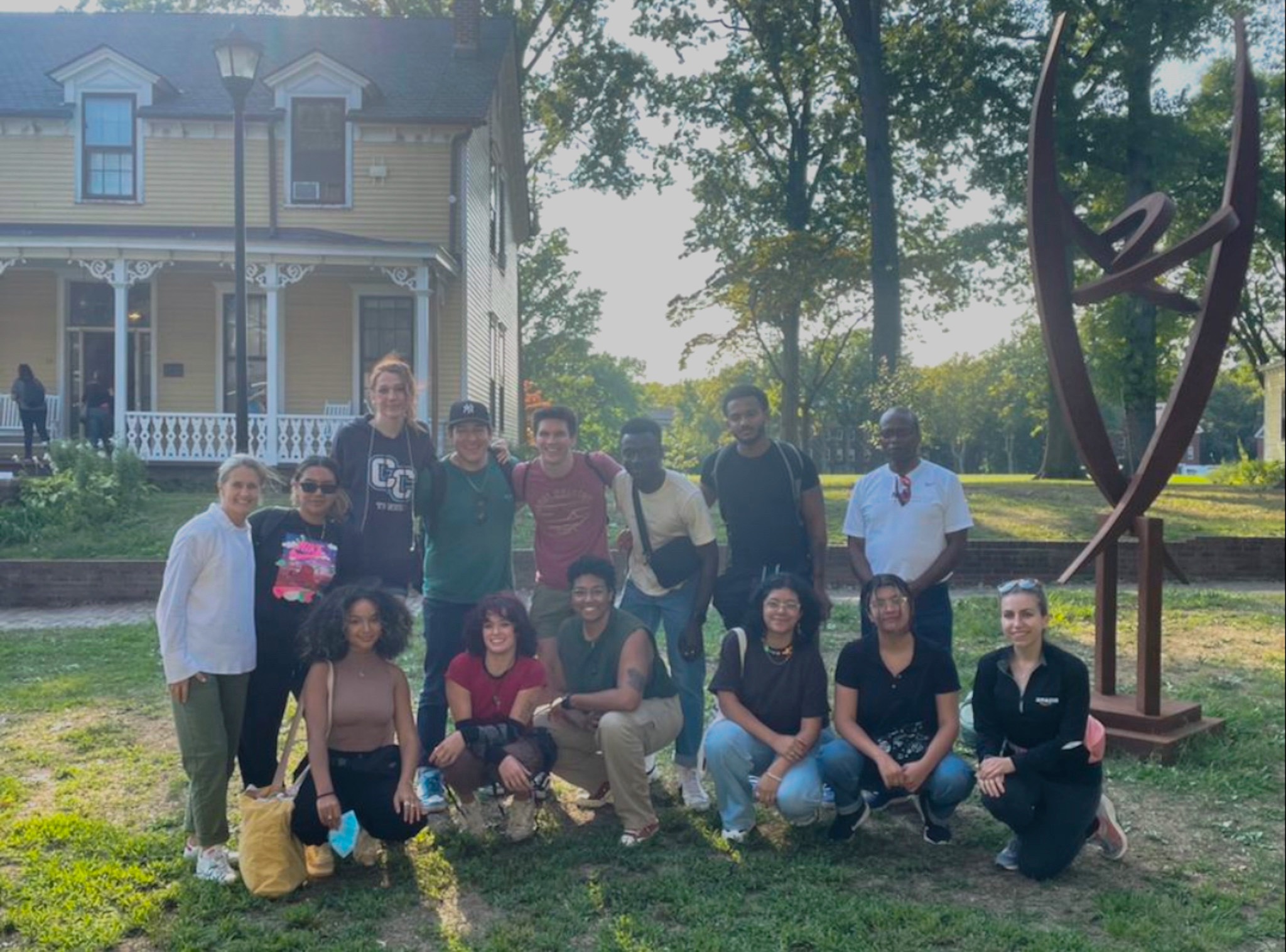 First-generation students standing and kneeling together in front of a yellow house and trees.