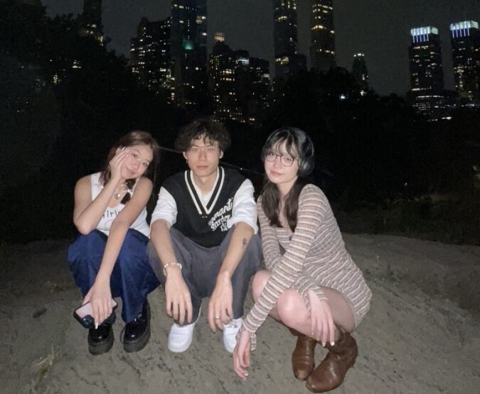 Three international students crouching in front of the New York City skyline at night.