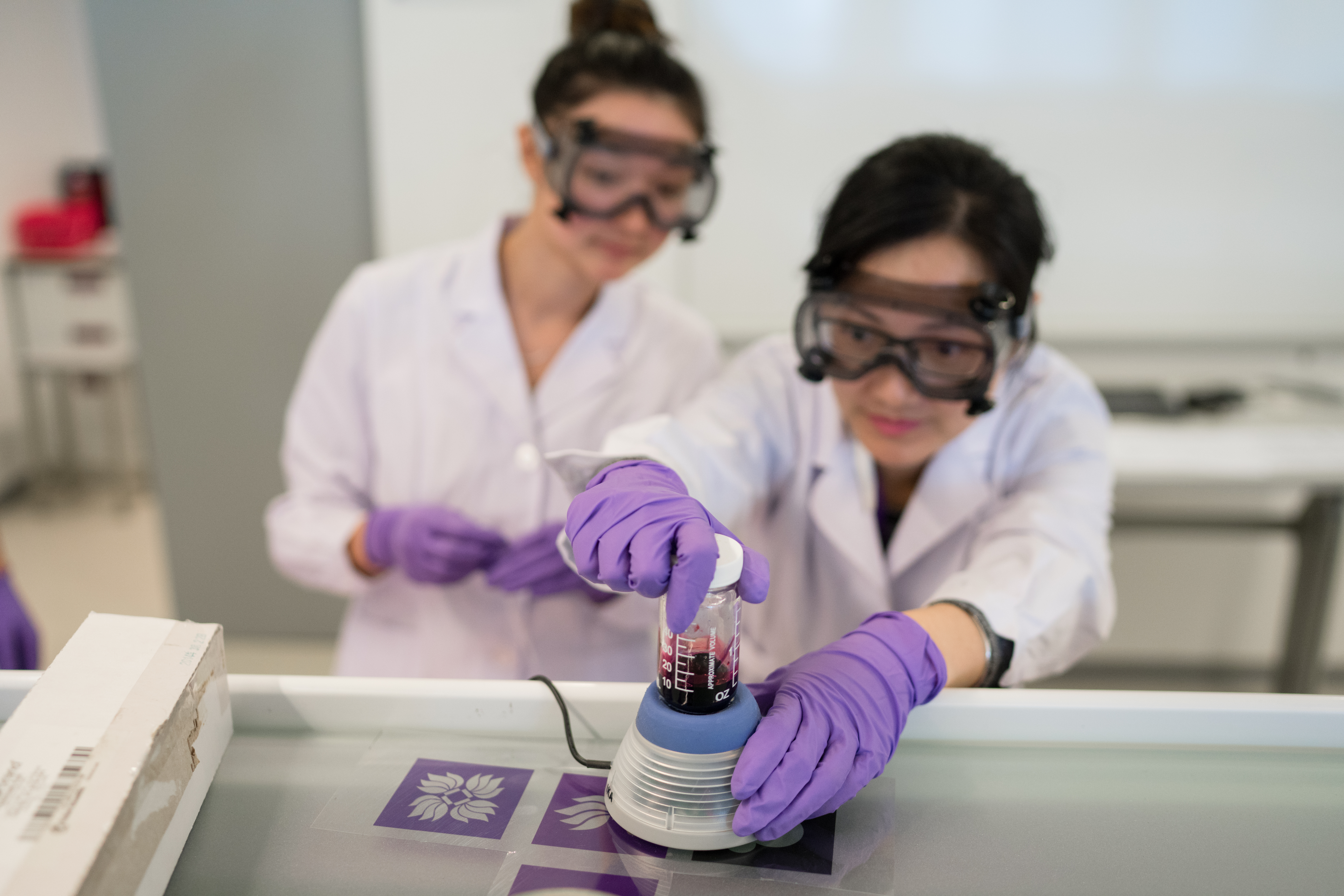 Two students wearing goggles and purple gloves work with equipment in an organic chemistry lab.
