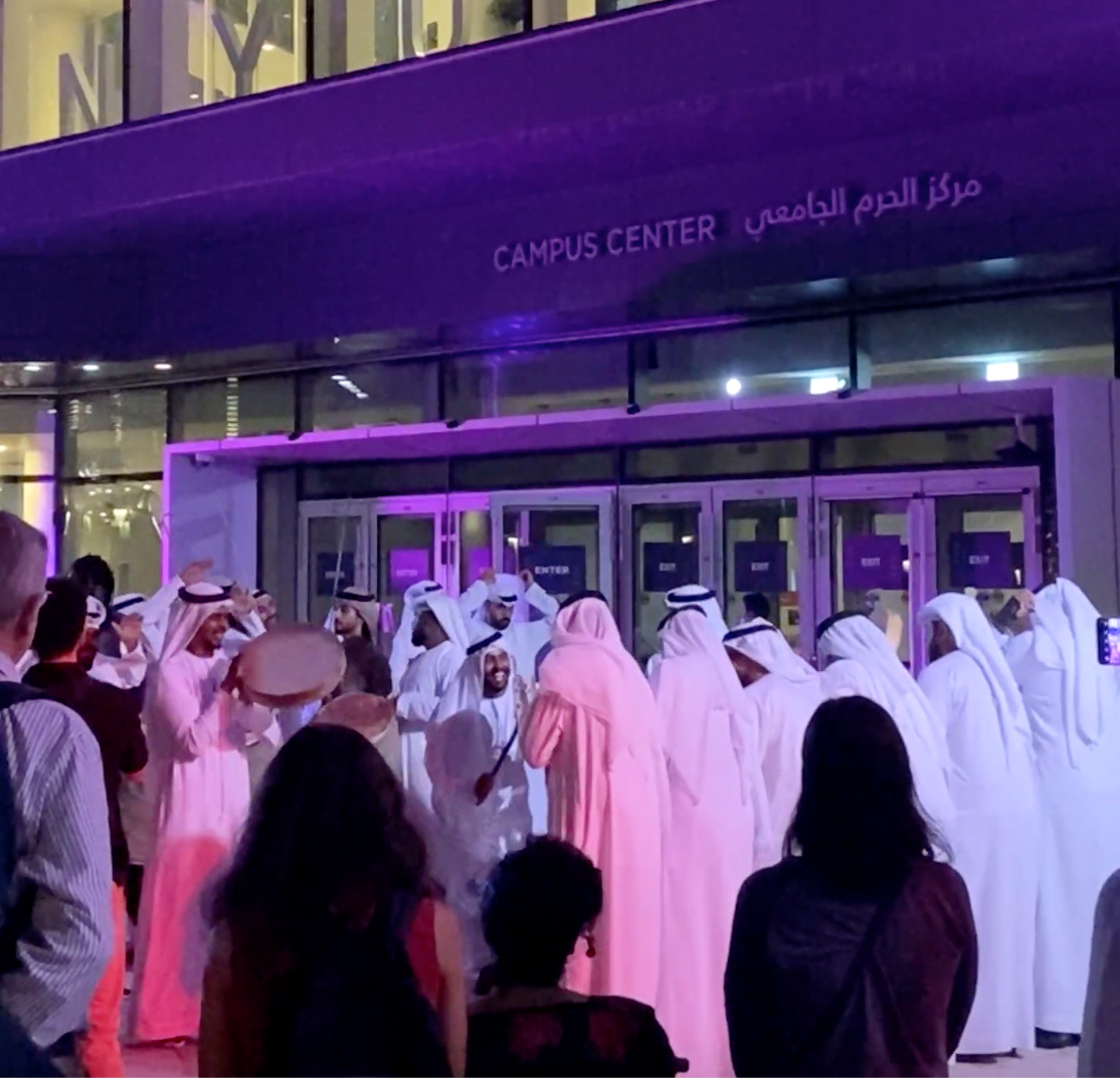 Several people standing outside of a violet-lit NYU Abu Dhabi campus building.