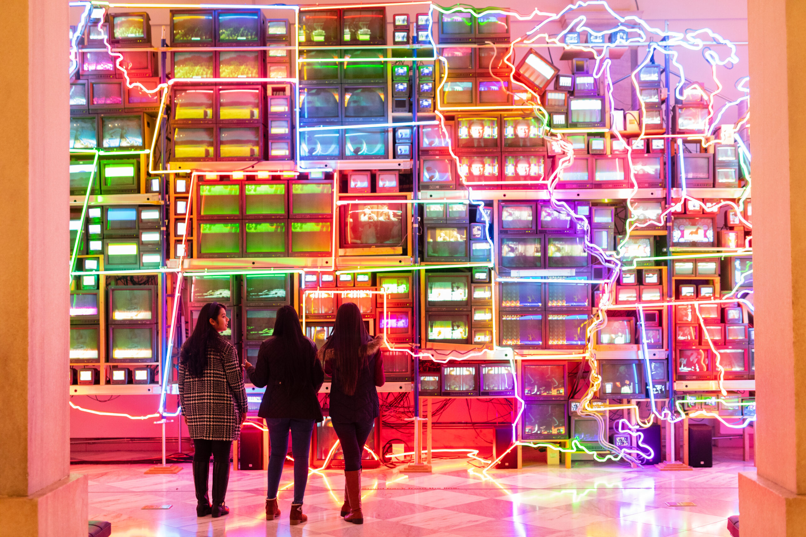 Three students standing in front of Nam June Paik’s “Electronic Superhighway: Continental U.S., Alaska, Hawaii,” a colorful light installation of 336 televisions, 50 DVD players, 3,750 feet of cable, and 575 feet of multicolored neon tubing.