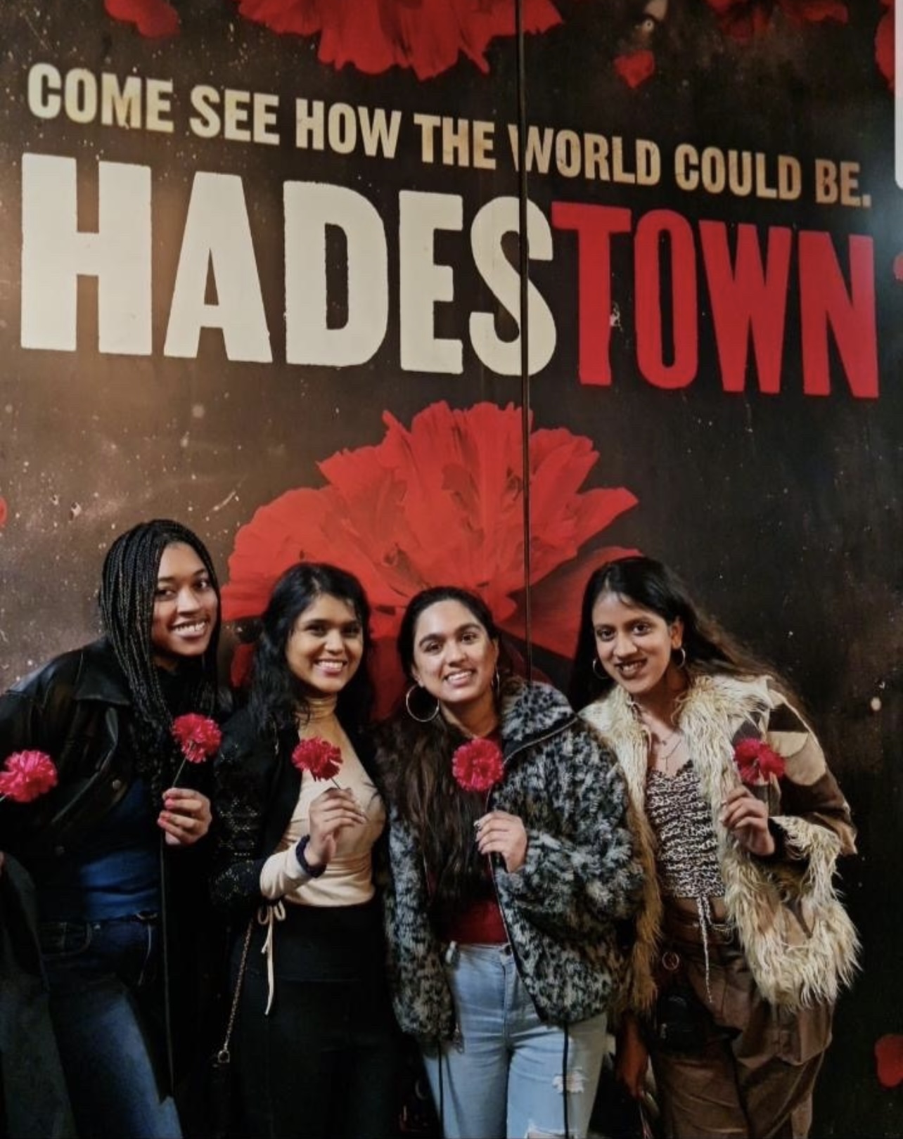 The author and some friends hold flowers and pose in front of a “Hadestown” poster.