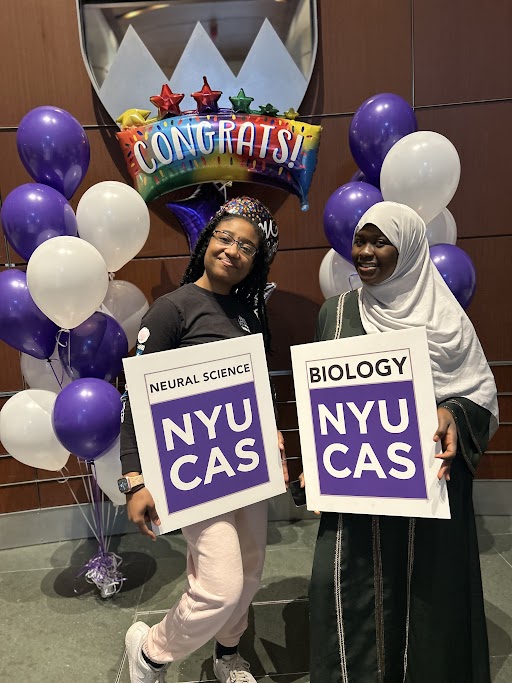 Two students holding signs with their declared majors, Neural Science and Biology, on them with violet and white balloons in the background.
