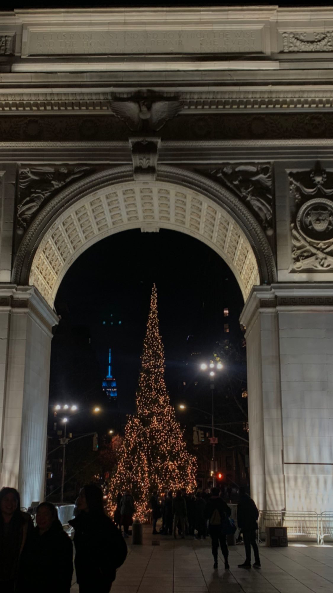 The Washington Square Arch at night with a view of the Empire State Building and a Christmas trees decorated in lights for the winter season.
