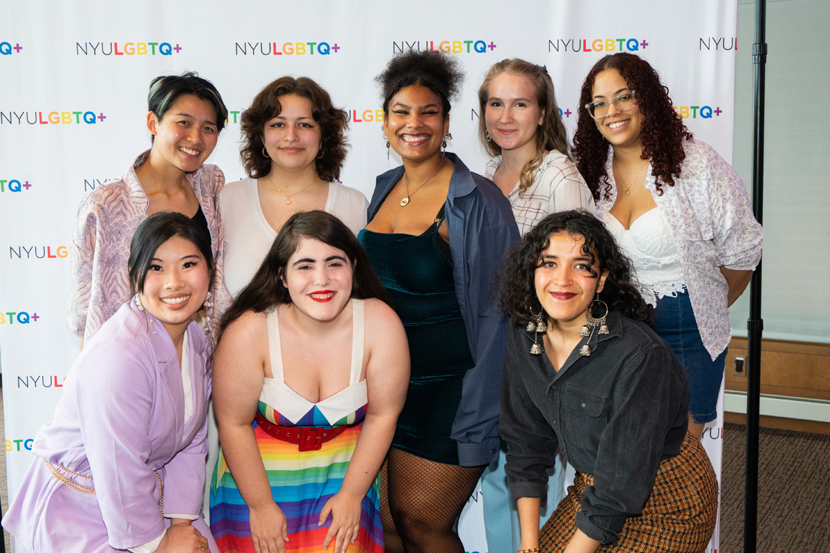 Eight members of CampGrrl pose together in front of a backdrop that reads, “NYULGBTQ+.”