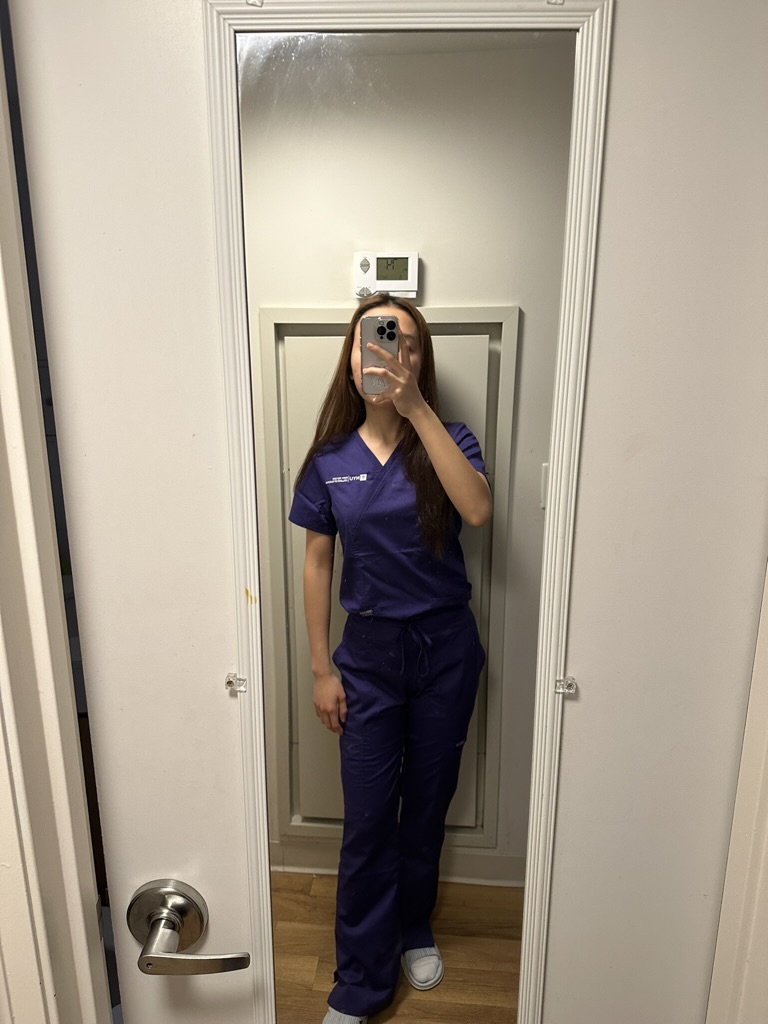 An NYU nursing student using a mirror while wearing scrubs and holding their cell phone in front of their face.