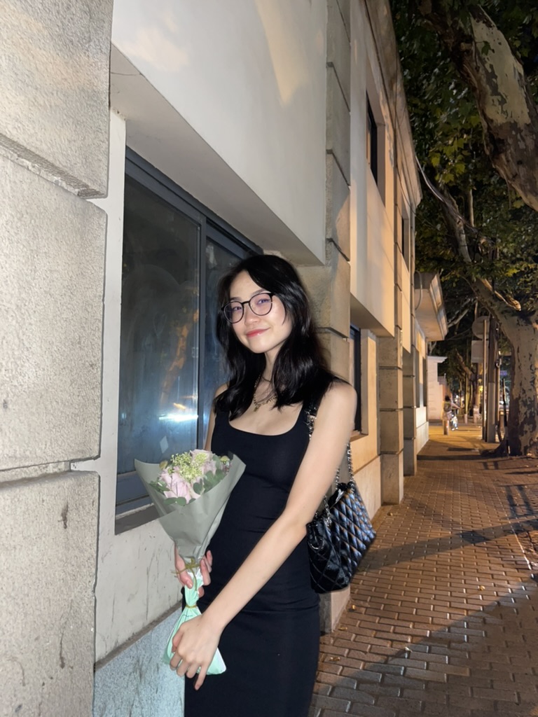 Yasmin, the author, stands in front of a building at night. She is smiling and holding a bouquet of flowers.