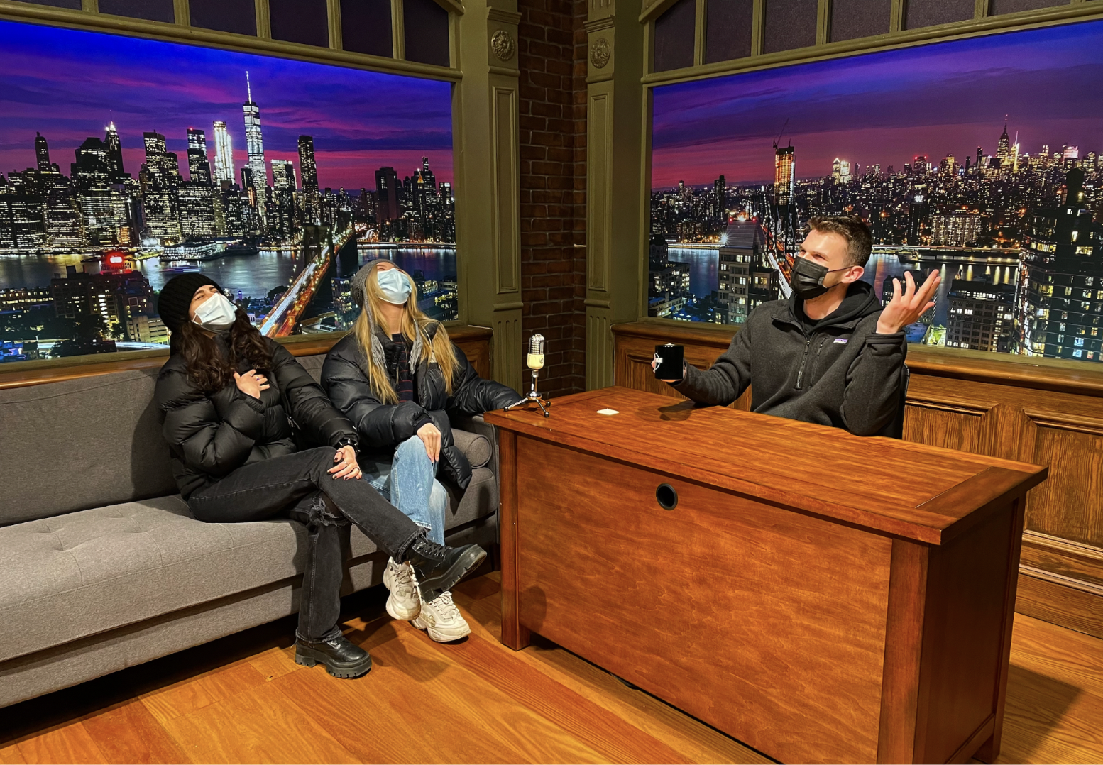 College Cohort students wearing masks. One student sits behind a late-night show host’s desk and the other two sit on the guest couch. All are laughing.
